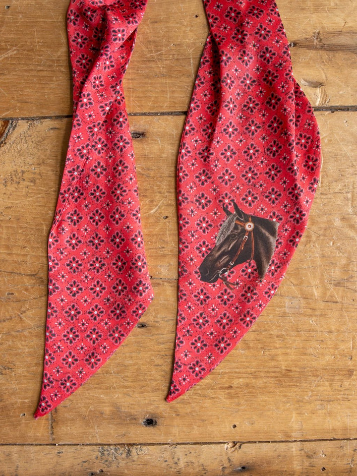 The Howdy Red Satin Tie Scarf by Savannah Sevens western life{&}style, featuring a horse head illustration on one end and a bold red Western pattern, is laid out on a wooden surface.