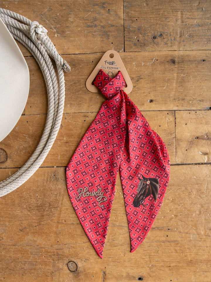 The Howdy Red Satin Tie Scarf by Savannah Sevens western life{&}style, showcasing a bold red pattern with a black horse illustration and the word "Howdy," is displayed on a wooden surface beside a coiled rope and a white hat.