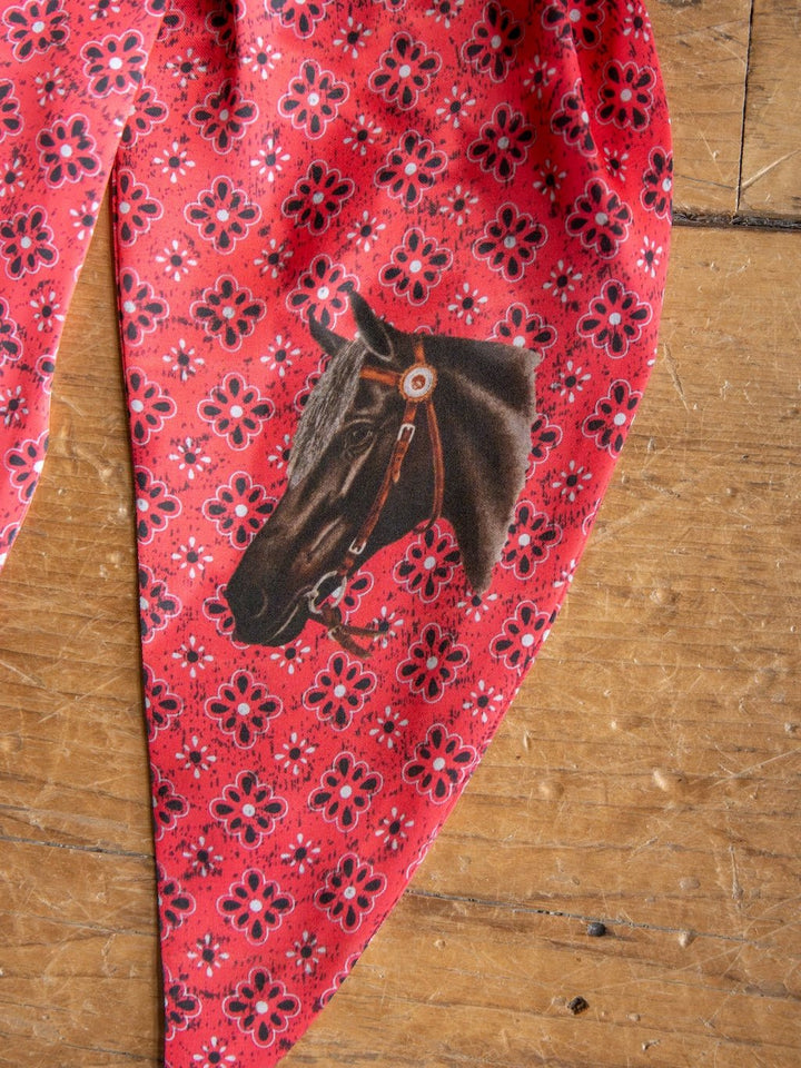 The Howdy Red Satin Tie Scarf by Savannah Sevens western life{&}style features a red patterned fabric adorned with black and pink floral designs and an illustrated horse’s head wearing a bridle. This western neck scarf is displayed on a wooden surface.