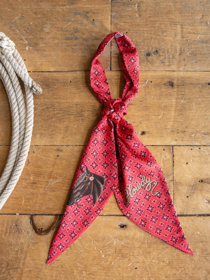 The Howdy Red Satin Tie Scarf by Savannah Sevens western life{&}style, featuring black and white patterns, a horse illustration, and the word "Howdy," is knotted on a rustic wooden surface beside a coiled rope.