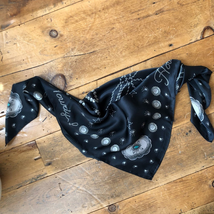 The Howdy Wild Rag {Noir} by Savannah Sevens western life{&}style—a black silk scarf with white and gray floral and circular patterns—is displayed folded in a triangle on a wooden floor, ends loosely tied for classic western style.