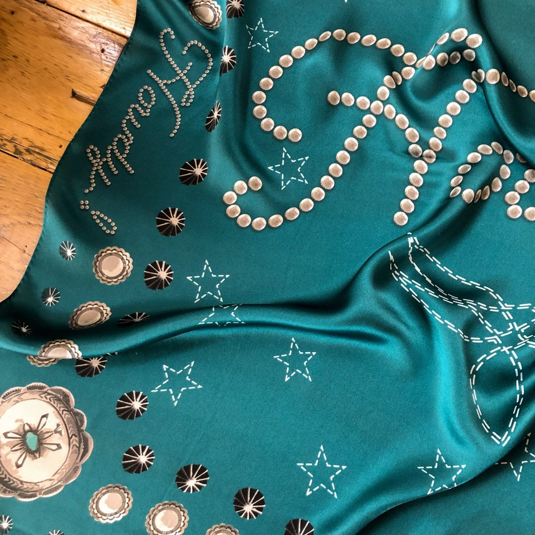 The Howdy Wild Rag {Teal} by Savannah Sevens western life{&}style, featuring embroidered white dotted stars, a framed bee, and cursive script with pearl-like dots, is draped elegantly on a wooden floor.