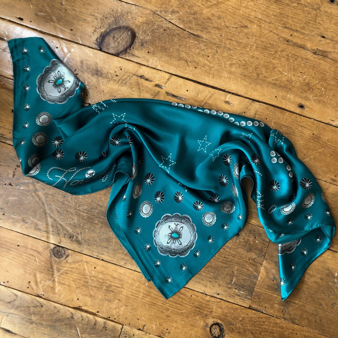 The Howdy Wild Rag {Teal} by Savannah Sevens western life{&}style features silver and white floral and star patterns, spread out with slight wrinkles on a rustic wooden floor—a chic western accessory.