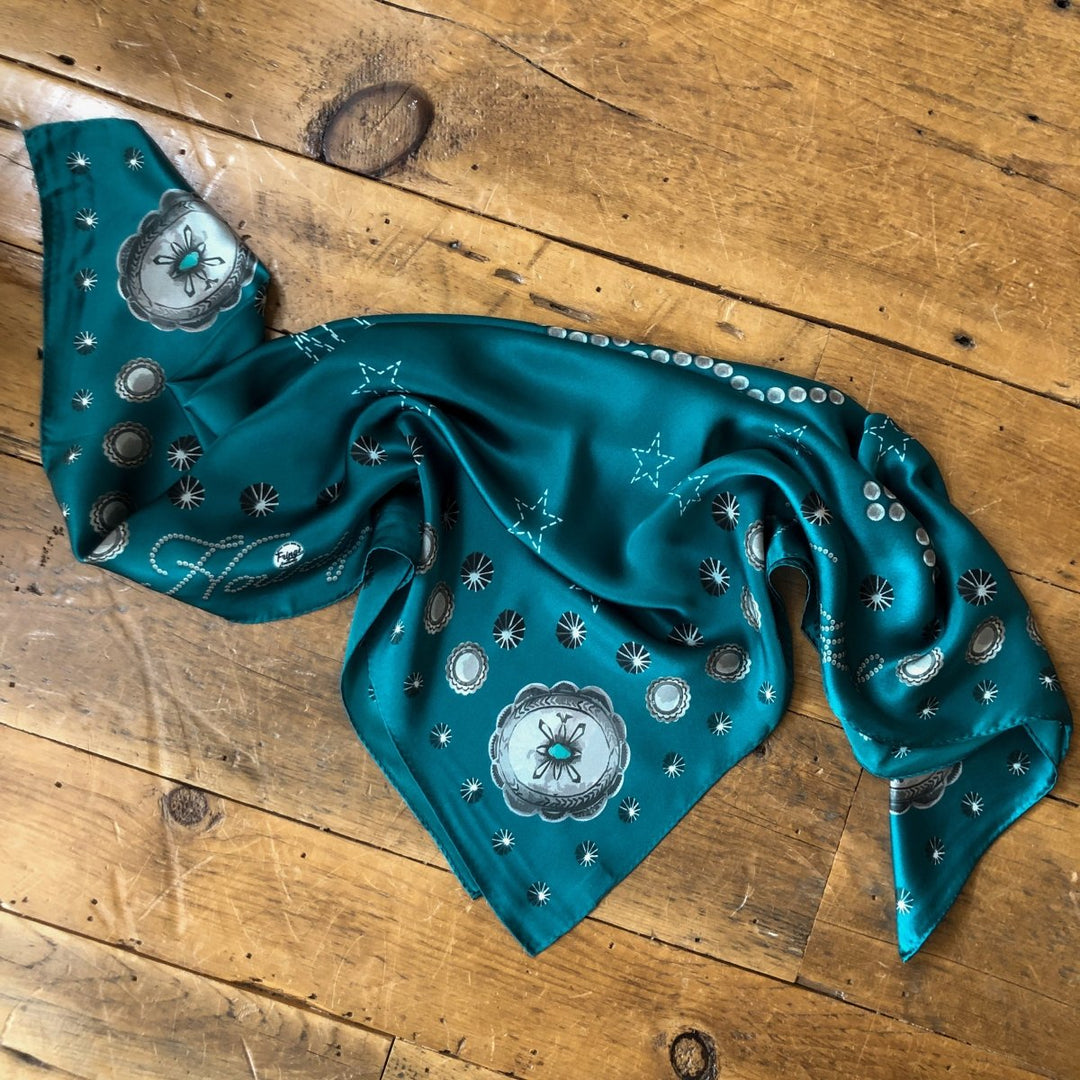 The Howdy Wild Rag {Teal} by Savannah Sevens western life{&}style features silver and white floral and star patterns, spread out with slight wrinkles on a rustic wooden floor—a chic western accessory.