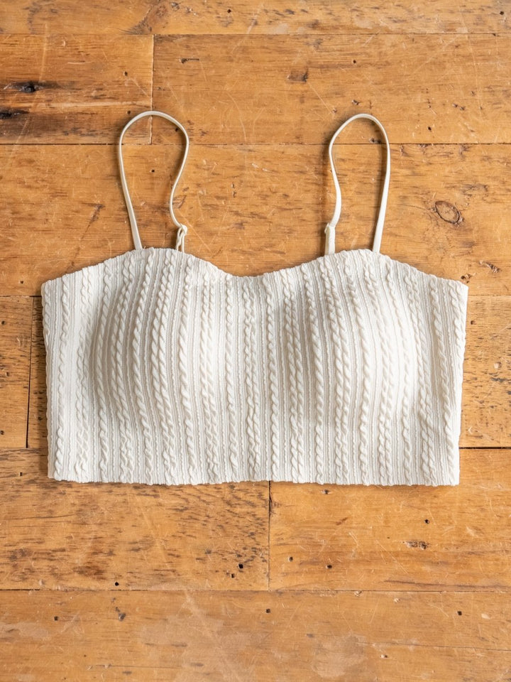 The Hoxie Bralette by Savannah Sevens western life{&}style features a cream cable knit, thin spaghetti straps, padded molded cups, and a vertical textured pattern, shown laid flat on a wooden surface.