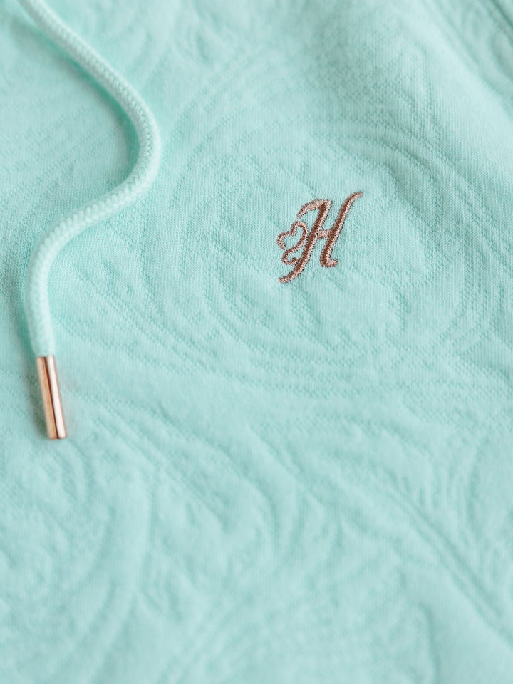 A close-up of The Hoyt hoodie by Savannah Sevens western life{&}style in seafoam, made from textured cotton blend with a drawstring featuring shiny metallic tips and a small brown embroidered H. Available in sizes S-XXL.
