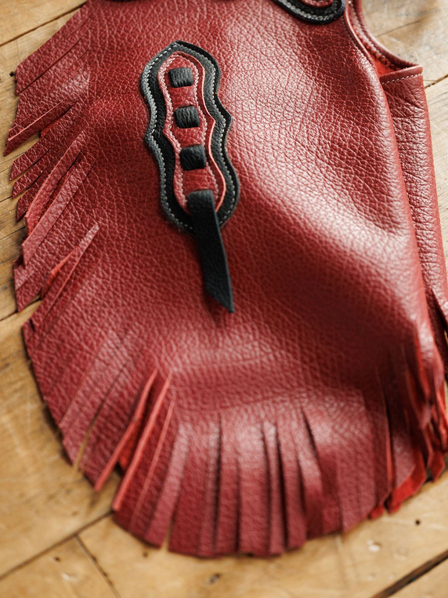 A close-up of Savannah Sevens western life{&}style’s Hulett Chinks {Toddler & Youth} in red leather with fringed edges and black stitched design, displayed on a wooden surface.