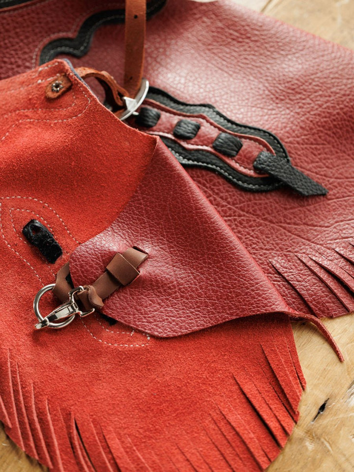Close-up of Hulett Chinks {Toddler & Youth} by Savannah Sevens western life{&}style—red and brown handmade leather with fringe, decorative stitching, metal buckles, loops, and adjustable waist on a wooden surface.