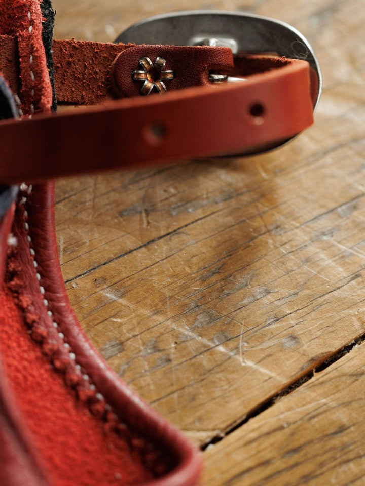 Close-up of Hulett Chinks {Toddler & Youth} by Savannah Sevens western life{&}style, showing red leather strap, metal hardware, detailed stitching, star-shaped rivet, and the textures of both premium leather and rustic wood.