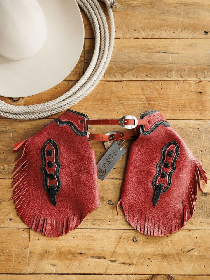 The Hulett Chinks {Toddler & Youth} by Savannah Sevens western life{&}style, featuring red leather with black detailing and fringe, lay on wood beside a light cowboy hat. Handmade with an adjustable waist and branded tag attached.