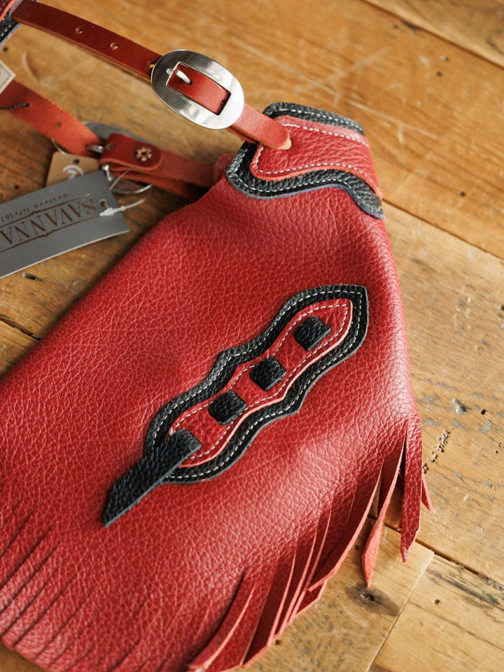 A close-up of Hulett Chinks {Toddler & Youth} by Savannah Sevens western life{&}style, featuring red leather, black and white stitching, black cutout details, fringe edges, and a silver buckle on a wooden surface.