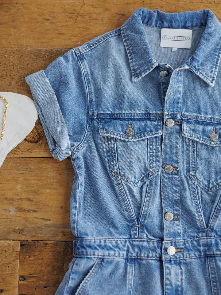 The Humboldt Denim Button Dress by Savannah Sevens western life{&}style, a short-sleeved western-style button-up with chest pockets, is displayed on a wooden surface beside a white fabric item with a gold chain.