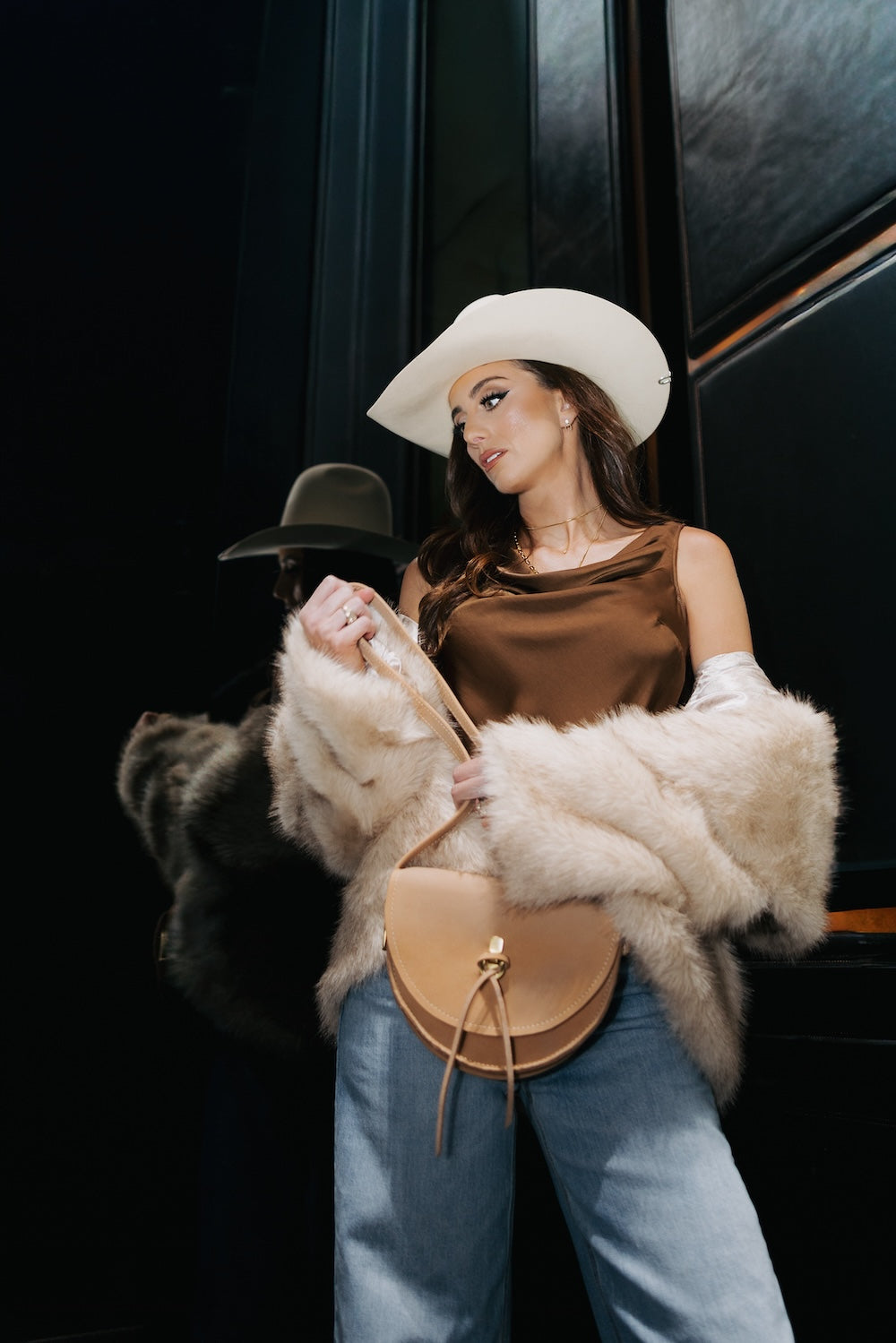 A woman in a white cowboy hat, brown satin top, light fur coat, blue jeans, and a tan crossbody bag stands confidently indoors, her mirror reflection behind her highlights her stylish presence.