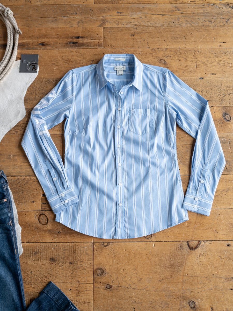 The Ingall by Savannah Sevens western life{&}style is a light blue, fitted long sleeve shirt with white vertical stripes, a chest pocket, and wrinkle resistance. Shown laid flat on a wooden floor with part of jeans and a white top nearby. Available S-XXL.