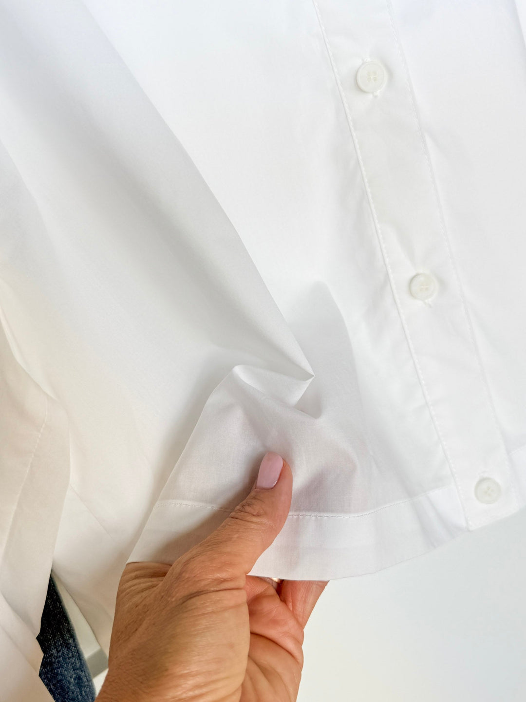 A hand with light pink nail polish pinches the hem of the Irving White Button Up Top by Savannah Sevens western life{&}style, showcasing its relaxed fit and soft texture.