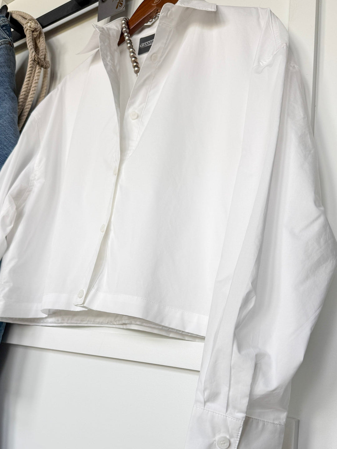 The Irving White Button Up Top by Savannah Sevens western life{&}style is displayed on a hanger against a white background, with part of a rope and blue fabric visible to the side.
