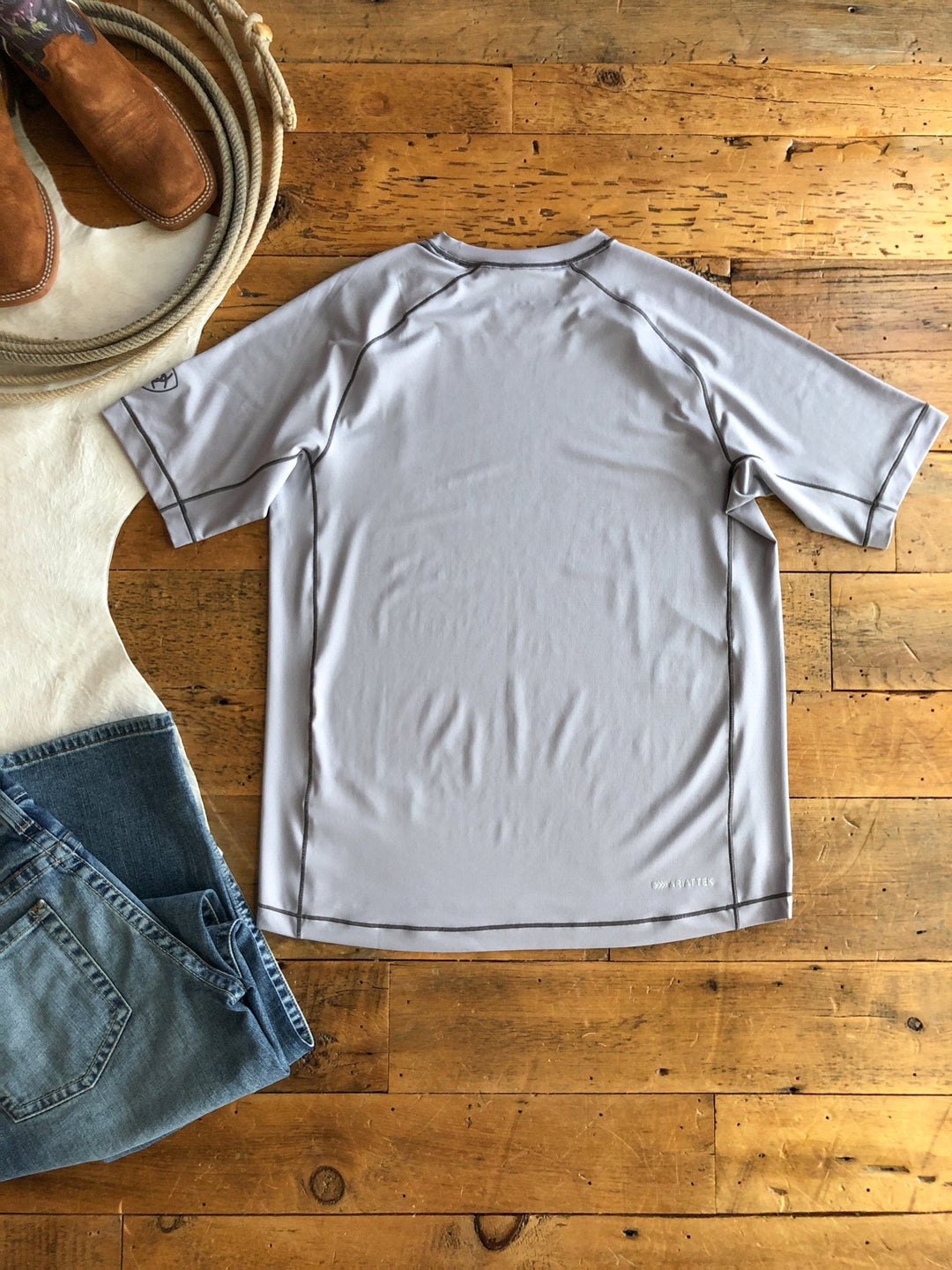 A light gray Jenks Ariat Men's Graphic Tee by Savannah Sevens western life{&}style is laid flat on a wooden floor, surrounded by blue jeans, cowboy boots, a coiled rope, and a white cowhide rug. The men's lightweight tee is shown from the back.