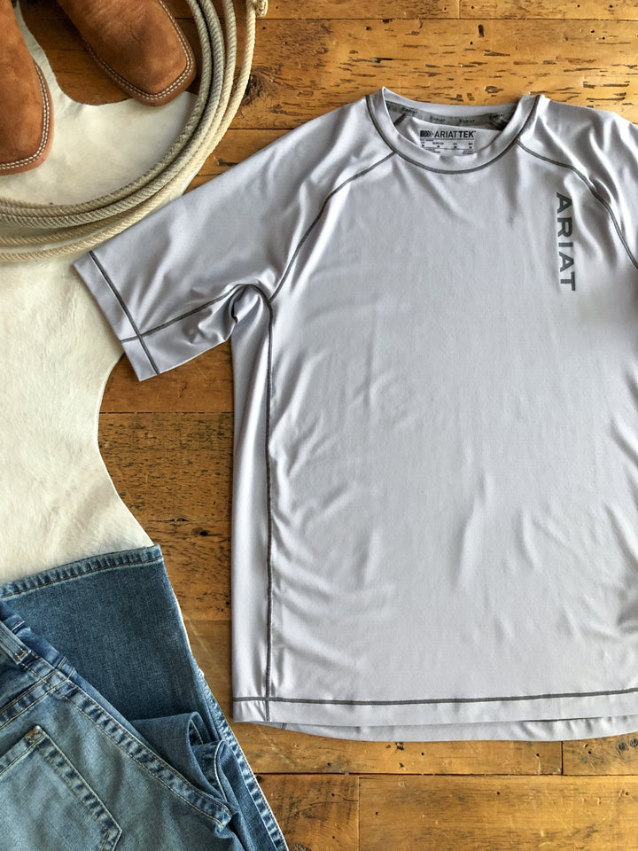 A Jenks Ariat Men's Graphic Tee by Savannah Sevens western life{&}style is laid flat on a wooden floor beside blue jeans, brown boots, and a white cowhide rug. This men's lightweight tee features moisture-wicking fabric for comfort.
