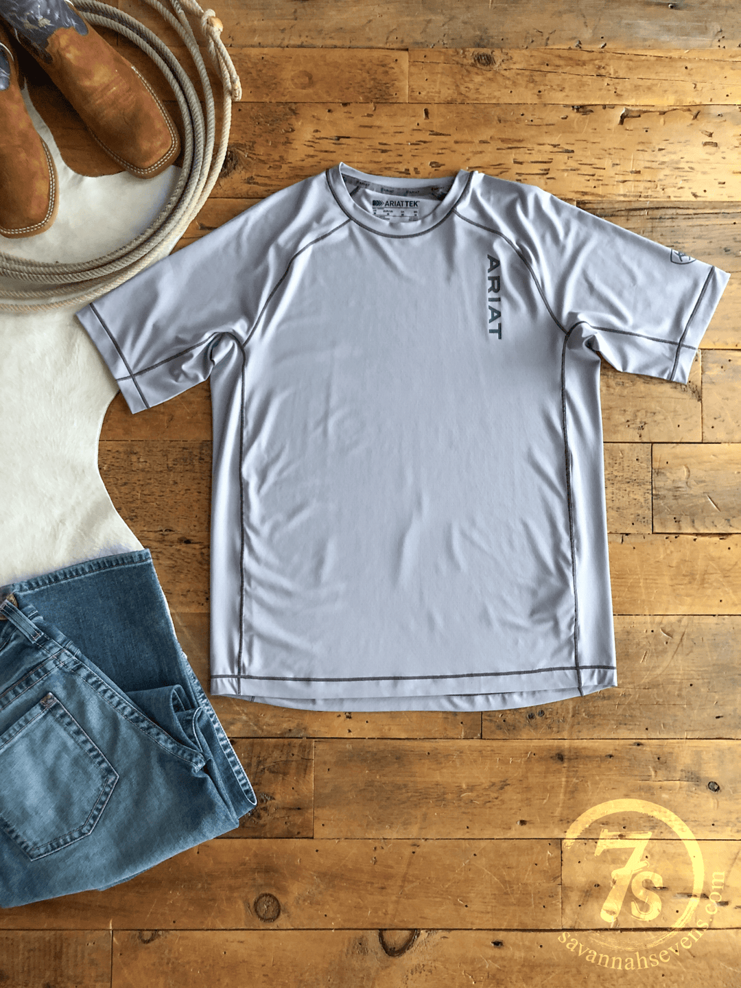 A light gray Jenks Ariat Men's Graphic Tee by Savannah Sevens western life{&}style, made with moisture-wicking fabric, is laid on a wooden floor beside blue jeans, brown cowboy boots, a coiled rope, and a white 7S logo rug.