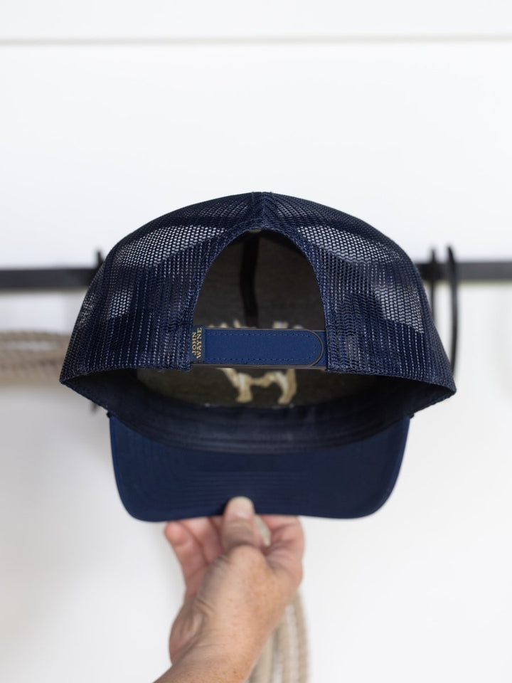 A hand holds a navy blue mesh John Wayne Cattle Co Cap by Savannah Sevens western life{&}style, showing the adjustable strap and back of the trucker hat against a white background.