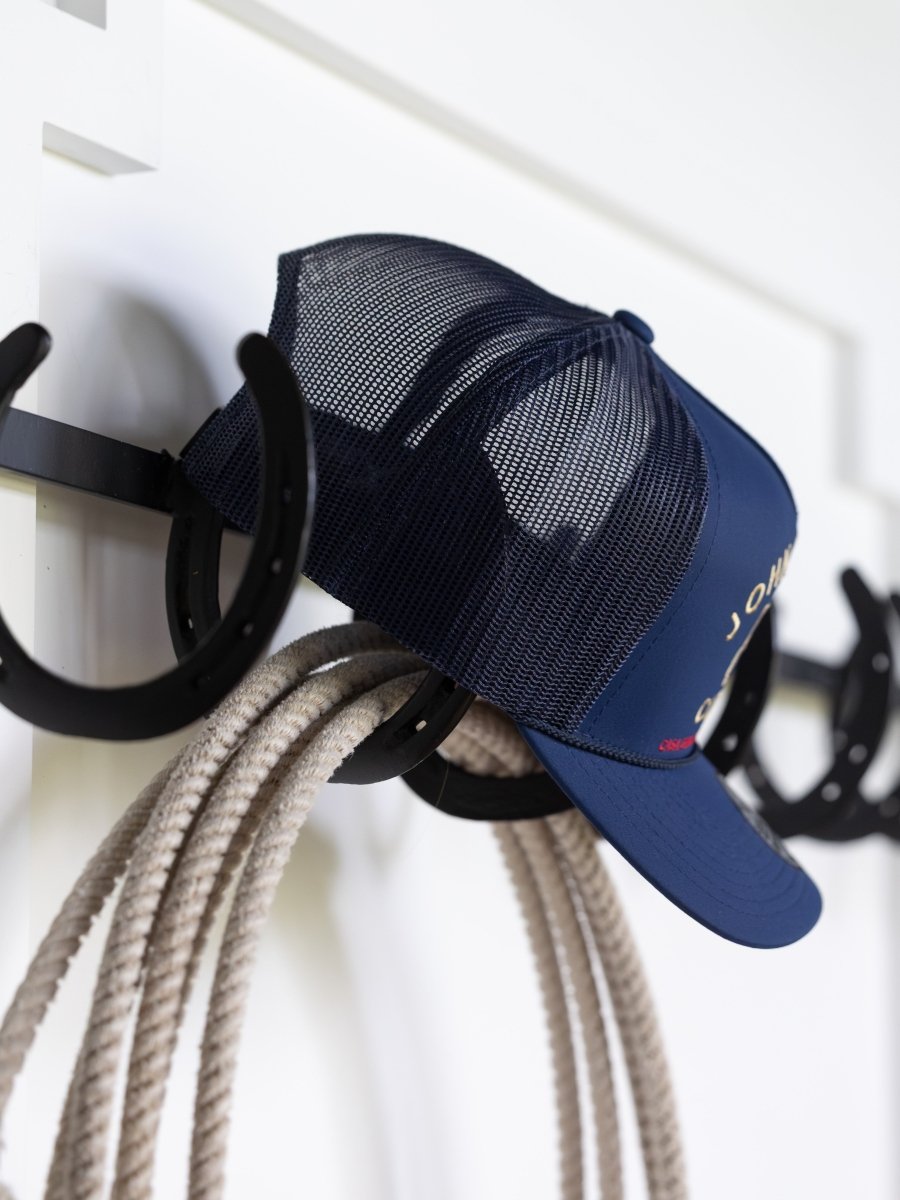 The John Wayne Cattle Co Cap by Savannah Sevens western life{&}style hangs on a black horseshoe-shaped hook with a coiled rope below, set against a white wall.