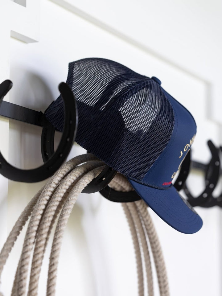 The John Wayne Cattle Co Cap by Savannah Sevens western life{&}style hangs on a black horseshoe-shaped hook with a coiled rope below, set against a white wall.