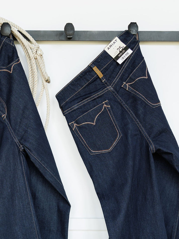 A pair of Ariat The Jolene High Rise Wide Leg Jeans with a 26M tag hangs on a black wall hook beside another pair of jeans and a coiled rope.