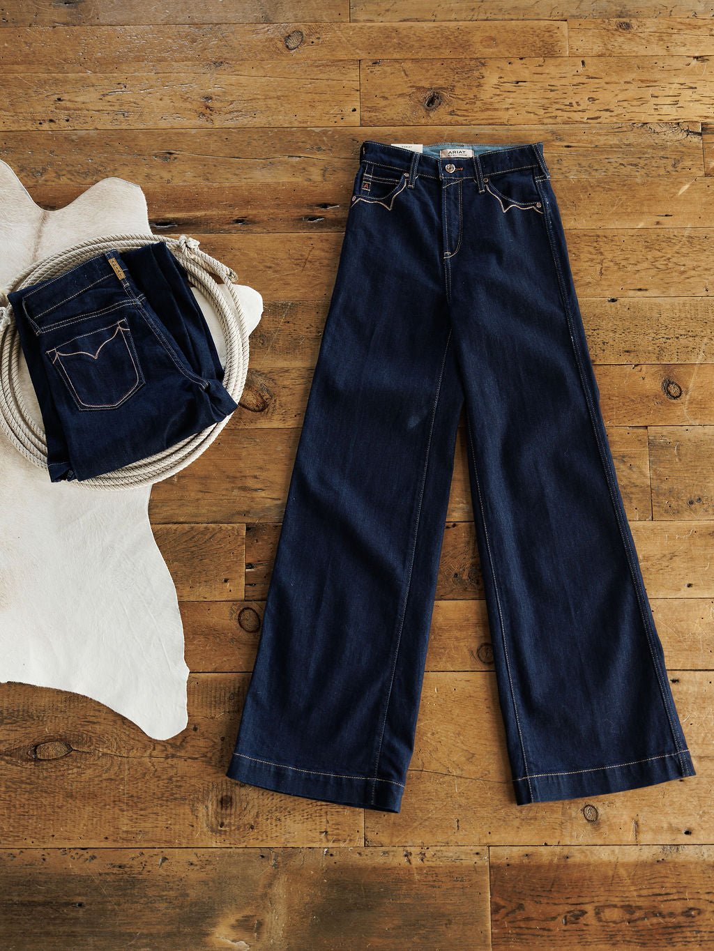A pair of Ariat The Jolene High Rise Wide Leg Jeans in dark wash laid flat next to a folded pair of Ariat jeans on a light animal hide, with a coiled rope nearby.
