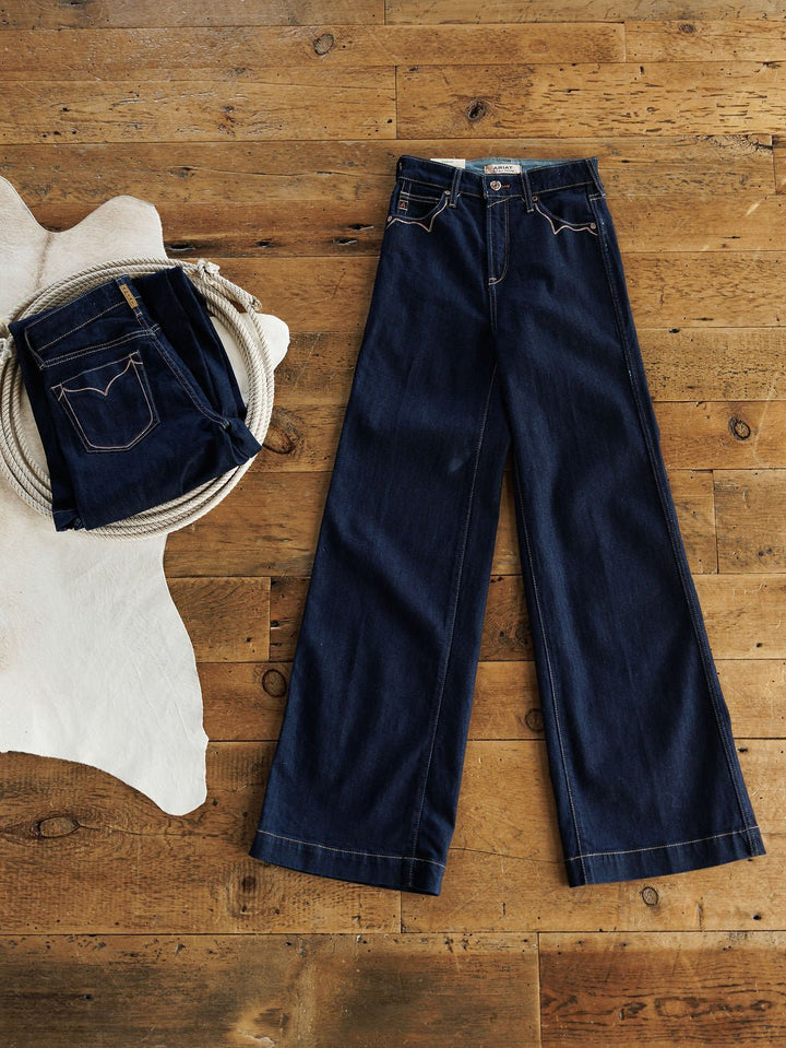 A pair of Ariat The Jolene High Rise Wide Leg Jeans in dark wash laid flat next to a folded pair of Ariat jeans on a light animal hide, with a coiled rope nearby.