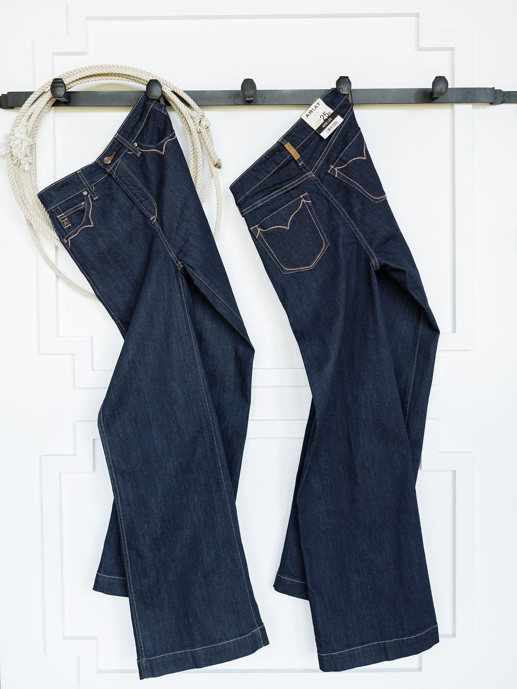 Two pairs of Ariat The Jolene High Rise Wide Leg Jeans hang side by side on a black rail, with a coiled rope draped over the left. The backdrop is a white wall featuring subtle panel detailing.