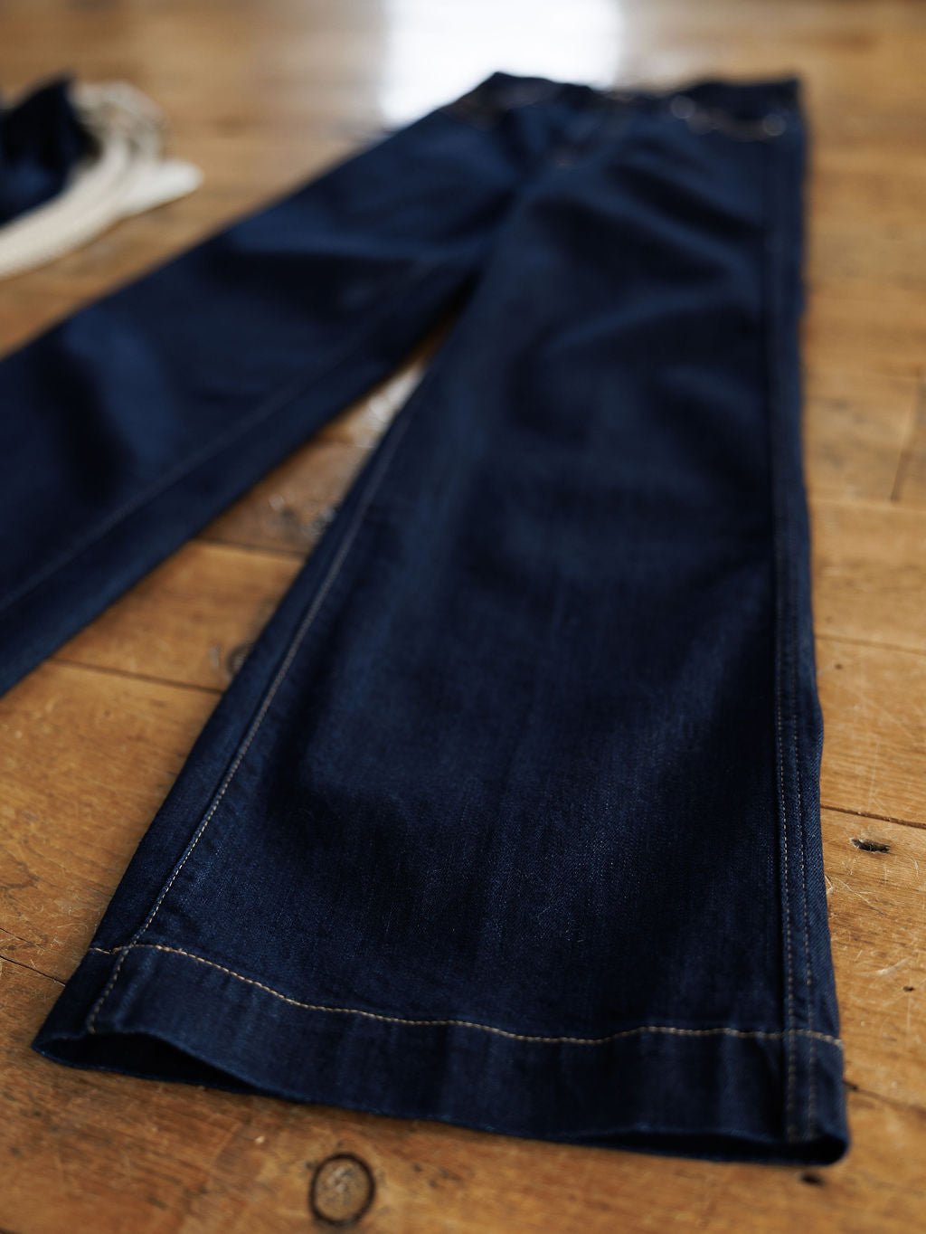 The Jolene High Rise Wide Leg Jean by Ariat, in dark wash denim, is laid flat on a wooden floor to showcase its ultra high rise fit and detailed stitching along the hem and seams.