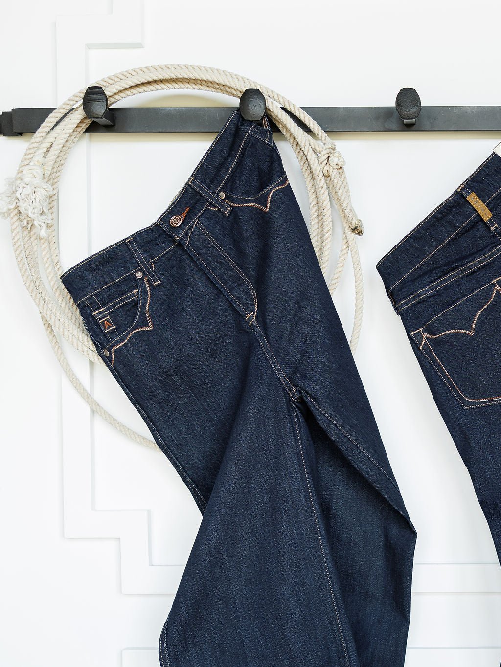 A pair of Ariat dark wash denim jeans hangs on a black hook rack with a coiled rope against a white wall. The Jolene High Rise Wide Leg Jean by Ariat is partially visible on the right.