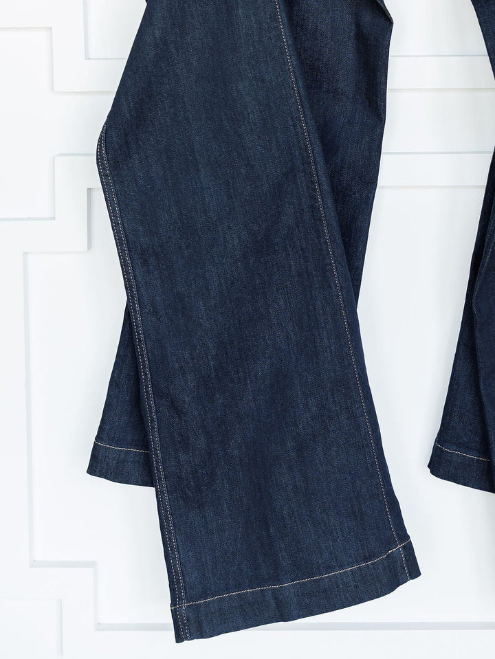The Jolene High Rise Wide Leg Jeans by Ariat, in dark wash denim with visible stitching, hang against a white geometric-patterned wall.