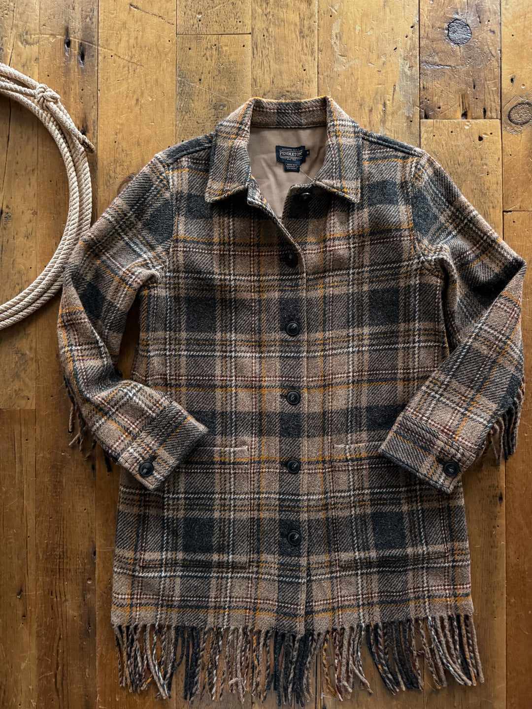 Savannah Sevens western life&style Jolene Plaid Fringe Wool Jacket laid flat on a wood floor, featuring a button-down front and fringe trim on the hem and cuffs.
