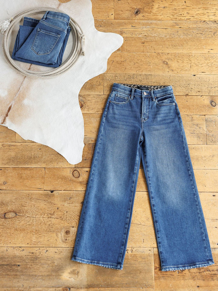 A pair of The Josey High Rise Wide Leg Crop Jeans by Savannah Sevens western life{&}style lies flat on a wooden floor, next to folded Risen Jeans in a woven basket atop a white cowhide rug.