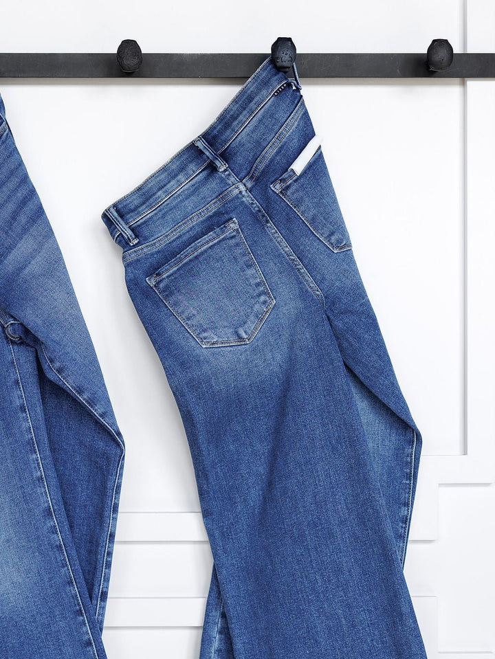 The Josey High Rise Wide Leg Crop Jean by Savannah Sevens western life{&}style hangs by the waistband on a black wall hook, showing a slight fade and back pocket. Another wide leg crop jean is partly seen on the left against a paneled white wall.