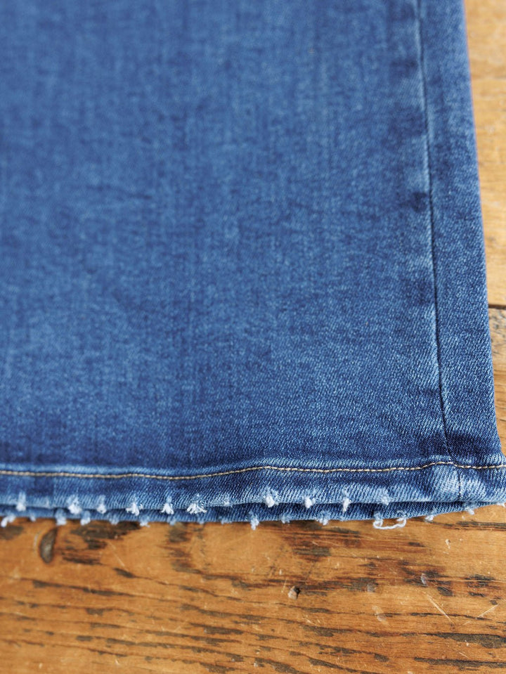 A close-up of the hem of Savannah Sevens western life{&}style's The Josey High Rise Wide Leg Crop Jean, featuring a blue wash and raw, frayed edge, lying flat on a wooden surface.