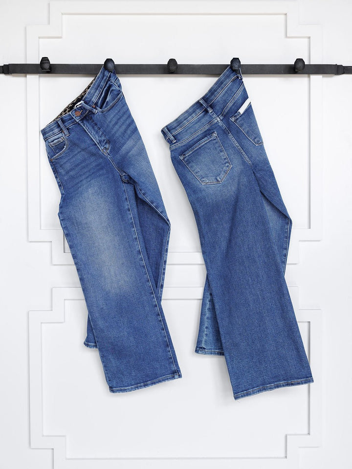 Two pairs of Savannah Sevens western life{&}style jeans, including The Josey High Rise Wide Leg Crop Jean, hang side by side on a black bar against a white paneled wall, legs slightly crossed to show front and back views.