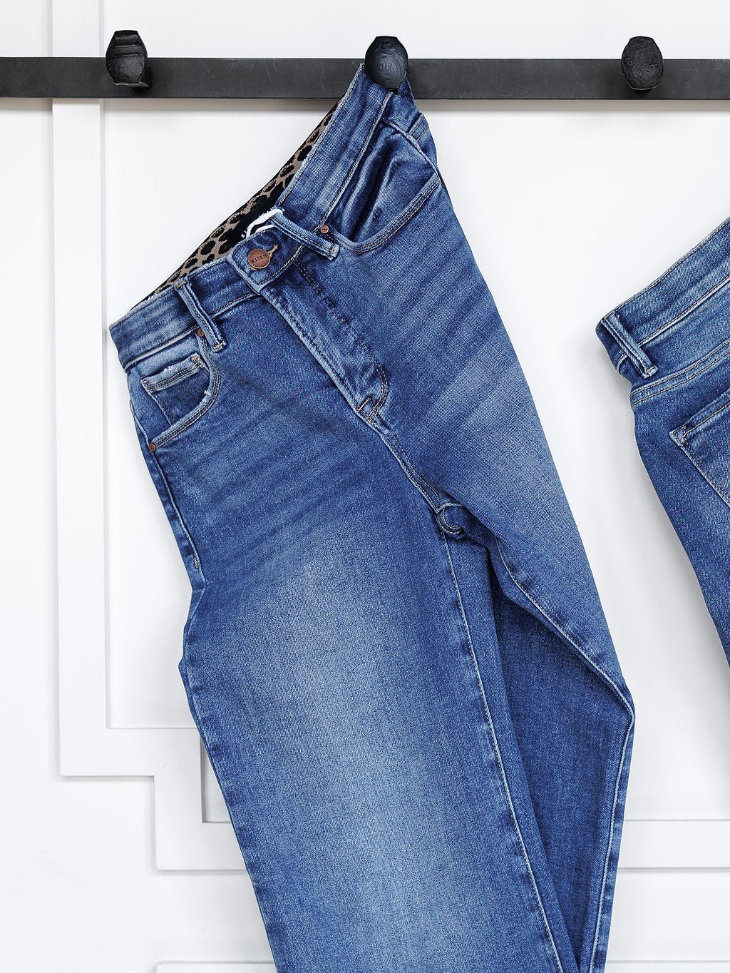 The Josey High Rise Wide Leg Crop Jean by Savannah Sevens western life{&}style in blue hangs by the belt loop from a black wall hook against a white paneled background.