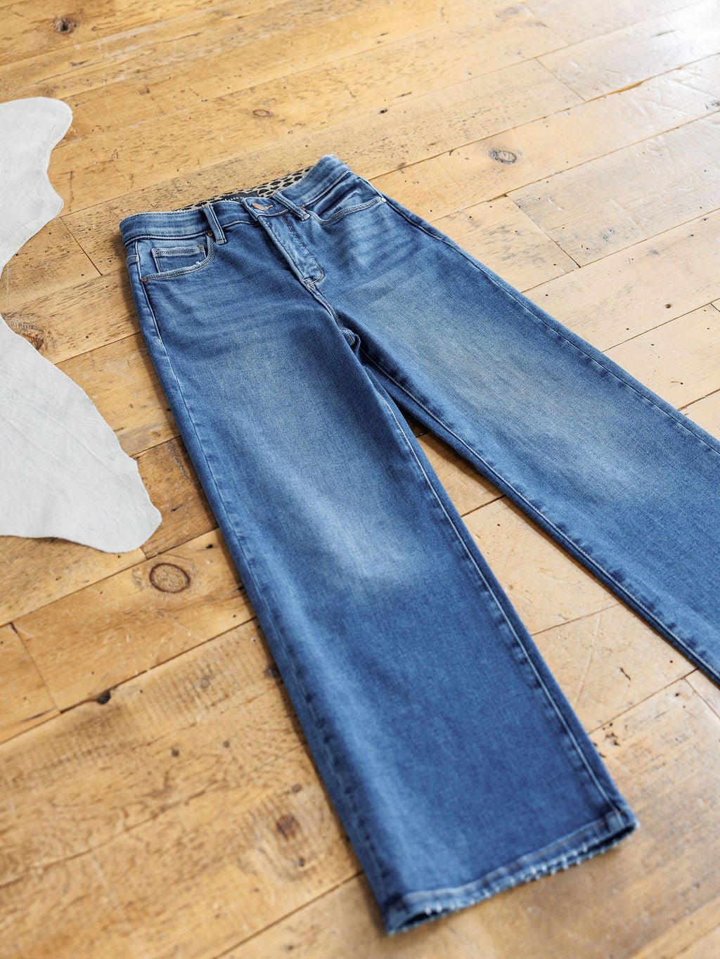 The Josey High Rise Wide Leg Crop Jean by Savannah Sevens western life{&}style is laid flat on a wooden floor, with a piece of white fabric partly visible on the left side of the image.