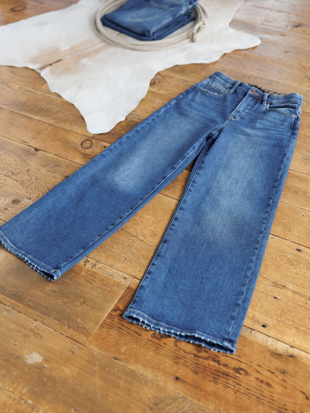 A pair of blue Risen Jeans with a raw hem and tummy control lies flat on a wooden floor. In the background, The Josey High Rise Wide Leg Crop Jean by Savannah Sevens western life{&}style is folded on a white animal hide rug.