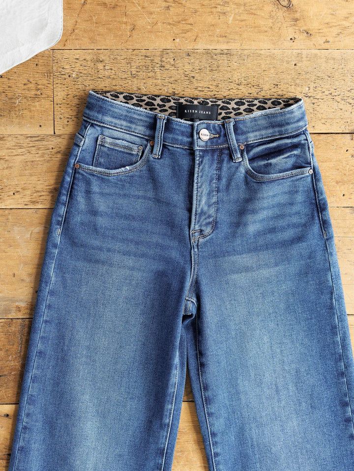 The Josey High Rise Wide Leg Crop Jean by Savannah Sevens western life{&}style features a blue wash and leopard print inner waistband, shown laid flat on a wooden surface.