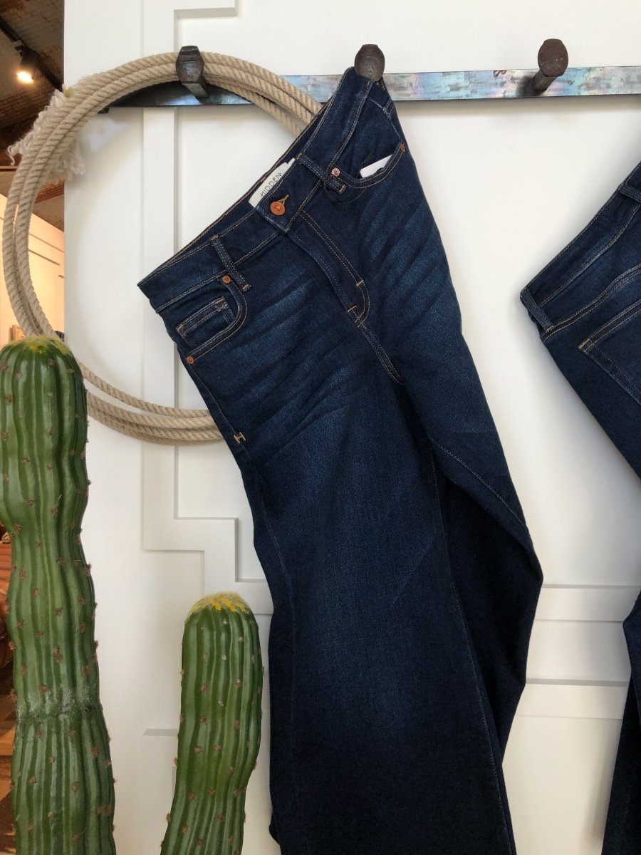 The Kade High Rise Wide Leg Crop Jean by Savannah Sevens western life{&}style in a dark wash hangs on a wall hook beside a coiled rope, with two tall green cacti in the foreground.