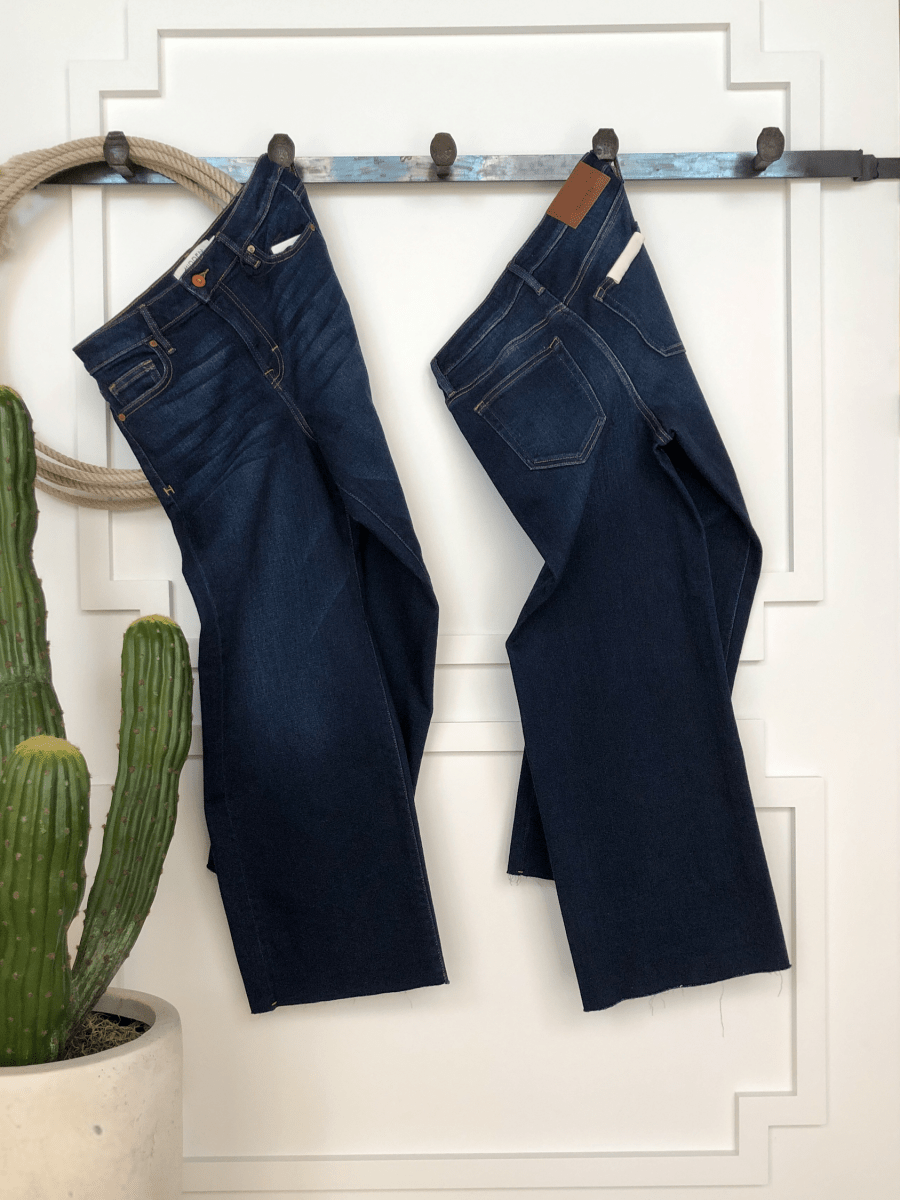 Two pairs of The Kade High Rise Wide Leg Crop Jean by Savannah Sevens western life{&}style, in a dark wash, hang side by side on hooks against a white wall. A tall green cactus in a white pot completes the cozy, rustic decor.