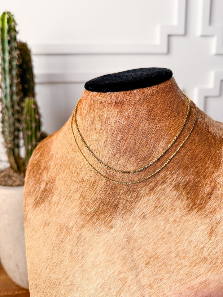 The Kahoka Necklace by Savannah Sevens western life{&}style, an 18K gold filled, hypoallergenic chain, is showcased on a brown and white cowhide jewelry bust with a potted cactus in the background against a white wall.