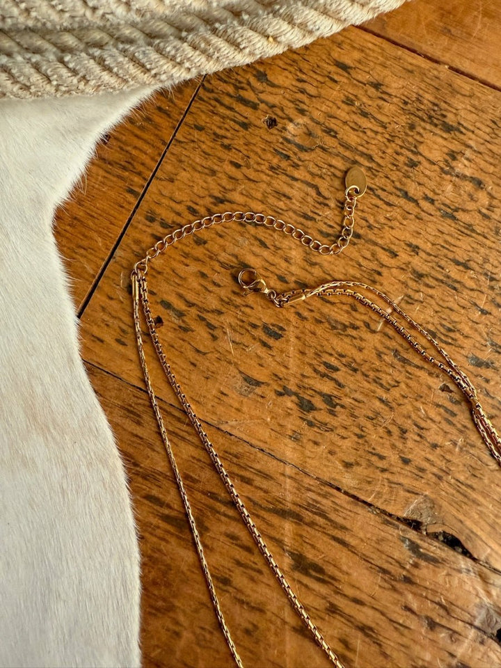 The Kahoka Necklace by Savannah Sevens western life{&}style is displayed on wood beside a white textured cowhide. Its 18K gold filled, hypoallergenic design combines elegance and comfort with striking contrasts in texture and tone.