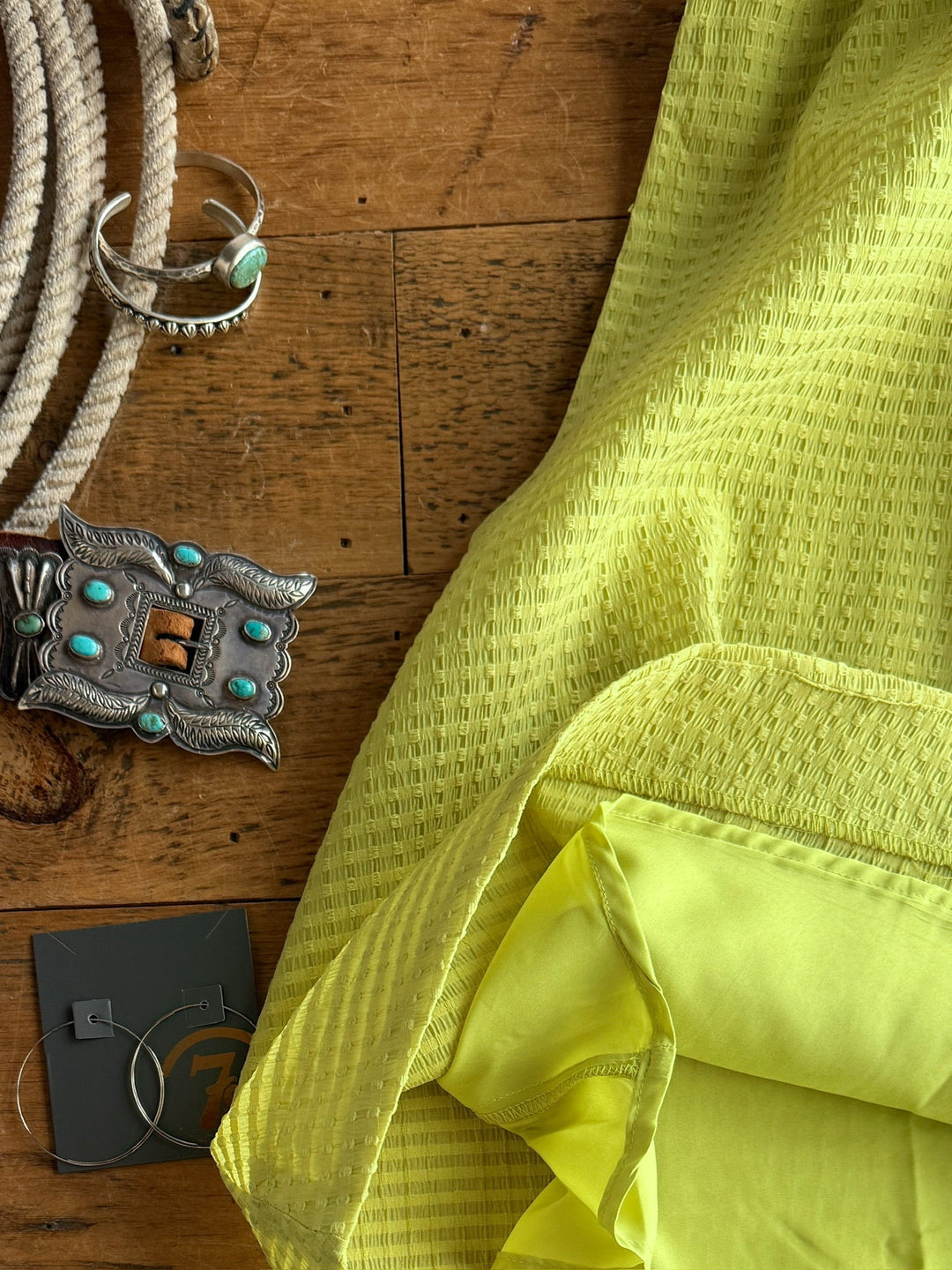 The Kalewa by Savannah Sevens western life{&}style—a yellow textured tri-blend with a ruched halter neckline—lies on wood next to a silver turquoise-studded belt buckle, turquoise bracelet, coiled rope, and hoop earrings.