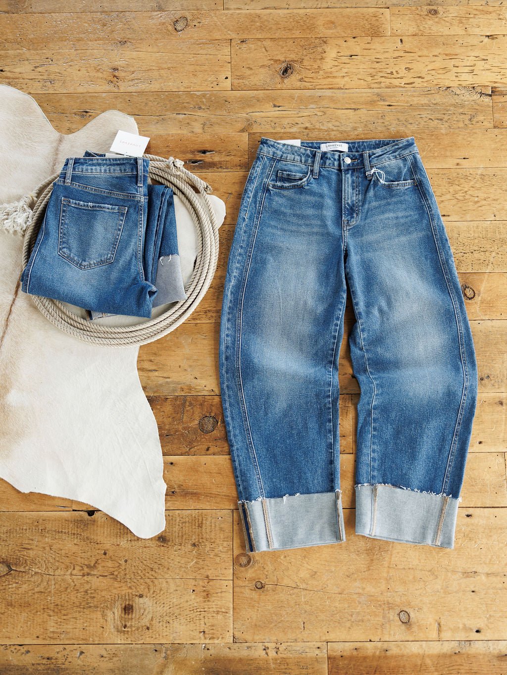 Two pairs of medium wash jeans from Savannah Sevens western life{&}style on a wooden floor: one folded with a tag on white hide, the other is The Kamden High Rise Barrel Jeans, laid flat with wide, cuffed hems.