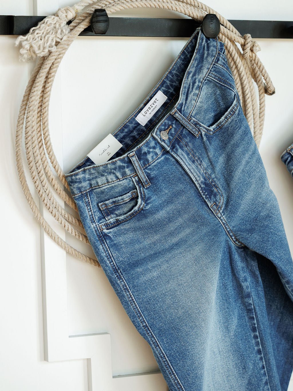 The Kamden High Rise Barrel Jeans by Savannah Sevens western life{&}style, in a medium wash with a visible white label and classic five-pocket design, hang over a decorative black hook with a coiled rope on a white wall.