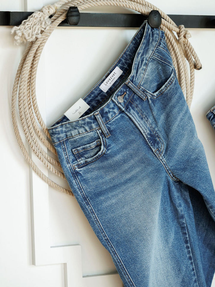 The Kamden High Rise Barrel Jeans by Savannah Sevens western life{&}style, in a medium wash with a visible white label and classic five-pocket design, hang over a decorative black hook with a coiled rope on a white wall.